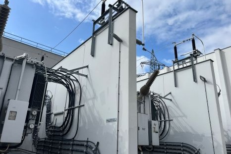 Stromtransformatoren mit Kabeln, unter blauem Himmel und Elektromasten.