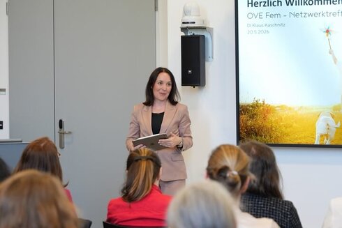 Eine Frau spricht bei einer Veranstaltung, im Hintergrund ein Willkommensschild.