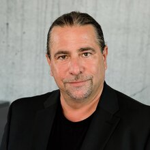 Ein Mann mit kurzen, dunklen Haaren in einem Anzug lächelt freundlich.
