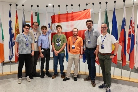 Gruppenfoto von sieben Personen vor einer Wand mit internationalen Flaggen.