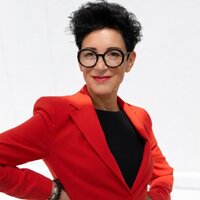 Frau in rotem Blazer lächelt, trägt große Brille und posiert selbstbewusst.