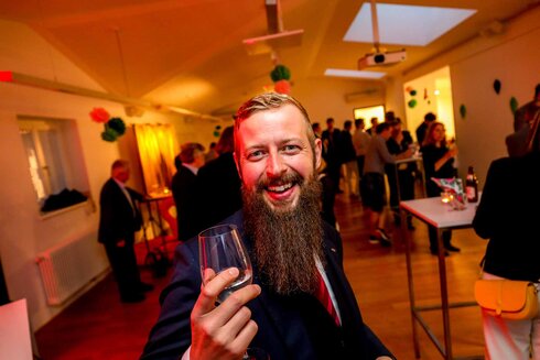 Mann mit Bart hebt ein Glas in festlicher Umgebung.