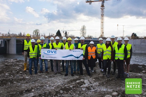 Gruppe von Bauarbeitern in Sicherheitskleidung hält ein Banner auf einer Baustelle.
