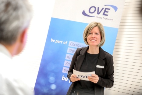 Eine lächelnde Frau hält ein Blatt Papier und spricht vor einem OVE-Stand.