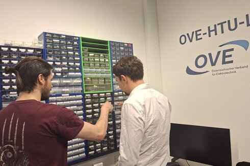 Zwei Personen betrachten eine Wand mit Werkzeug- und Elektronikbehältern.