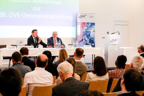 Zwei Redner auf einer Bühne bei der OVE-Generalversammlung. Publikum in Sicht.