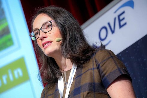 Eine Frau spricht auf einer Bühne mit Mikrofon und Brille.