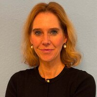 Frau mit blonden Locken, schwarzen Pullover und verschränkten Armen vor blauem Hintergrund.