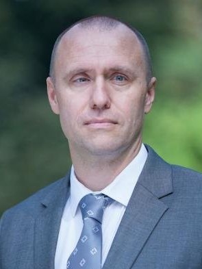 Ein Mann in Anzug und Krawatte, mit neutralem Gesichtsausdruck.