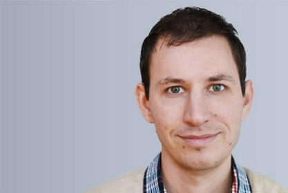Ein Mann mit braunen Haaren und blauem Hemd lächelt in die Kamera.