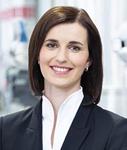 Lächelnde Frau in Anzug, vor einem modernen, industriellen Hintergrund.