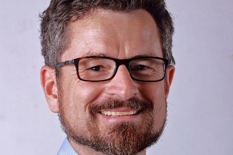 Porträt eines lächelnden Mannes mit Brille und Bart vor neutralem Hintergrund.